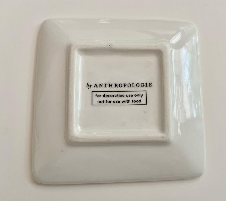 May include: A square, white Anthropologie decorative dish. The dish has rounded corners and a slightly raised square centre. Text on the bottom reads "by ANTHROPOLOGIE" and "for decorative use only not for use with food."