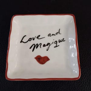 Op de afbeelding: Vierkante witte keramische schaal met een rode rand. De schaal heeft de woorden "Love and Magique" in zwart schrift, samen met een rode lip-afbeelding.