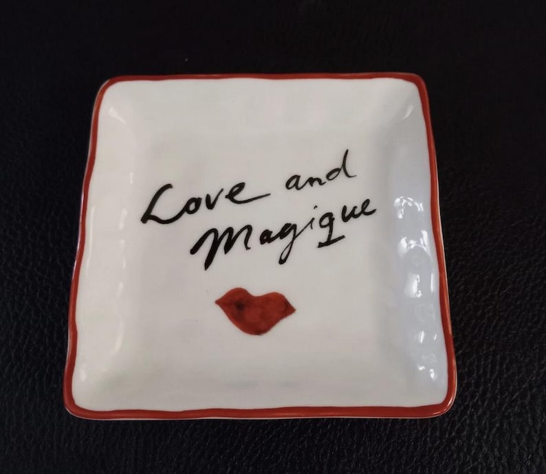 Op de afbeelding: Vierkante witte keramische schaal met een rode rand. De schaal heeft de woorden "Love and Magique" in zwart schrift, samen met een rode lip-afbeelding.