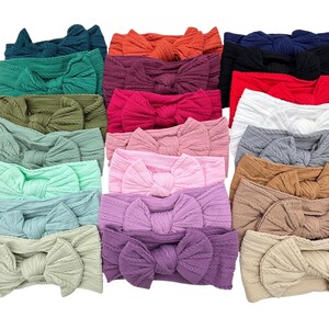 May include: A collection of colorful baby headbands with large bow accents. The headbands are in various shades, including teal, orange, burgundy, pink, white, gray, and tan. The bows have a textured, knitted appearance.