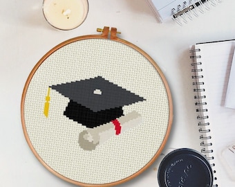 Peut inclure: Un cerceau de broderie blanc avec un motif de point de croix représentant un bonnet de diplômé et un diplôme. Le motif est en noir, gris, rouge et jaune.