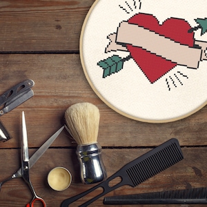 Puede incluir: Una mesa de madera con una variedad de herramientas de aseo, que incluyen un cepillo de afeitar, un peine, tijeras y una navaja. También hay un pequeño recipiente de crema de afeitar y un diseño de punto de cruz enmarcado de un corazón con una flecha que lo atraviesa.