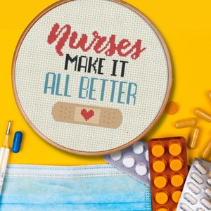 May include: A white embroidery hoop with the text "Nurses Make It All Better" in red and blue lettering. A red heart is on a brown bandage in the center of the hoop.