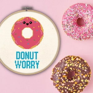 May include: A white embroidery hoop with a pink donut with colorful sprinkles and a smiling face. The text "DONUT WORRY" is written in blue below the donut.