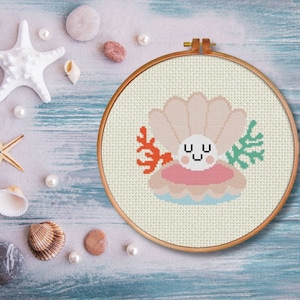 May include: A cross-stitch embroidery hoop with a white background featuring a pink and white seashell with a smiling face. The seashell is surrounded by coral and seaweed.