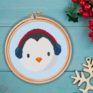 May include: A cross-stitch embroidery hoop with a penguin wearing red and blue headphones. The penguin is white with a black head and orange beak. The background is light blue.