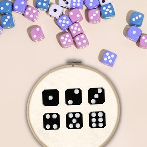 May include: A white embroidery hoop with black dice designs on a white fabric. The dice show the numbers one through six. The hoop is surrounded by blue and pink dice.