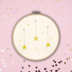 May include: A white embroidery hoop with a cross-stitch design of three yellow stars hanging from strings. The stars are outlined in black and have a pixelated appearance. The hoop is on a pink background with scattered gold and silver stars.