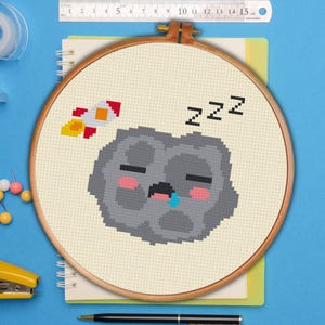 May include: A cross-stitch hoop showing a sleeping gray asteroid with pink cheeks and a blue tear, and a red and yellow rocket. The word "ZZZ" is stitched above the asteroid, indicating sleep. The hoop is set on a cream-coloured fabric.