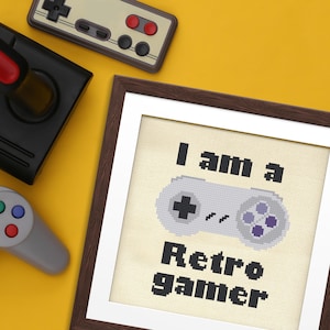 May include: A framed cross-stitch art piece with the text "I am a Retro gamer" and a pixelated game controller. The image is on a yellow background with retro game controllers.
