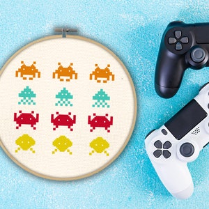 Op de afbeelding: Een witte borduurring met een kruissteekpatroon van kleurrijke gepixelde space invaders. De ring is op een blauwe achtergrond met twee videogamecontrollers.