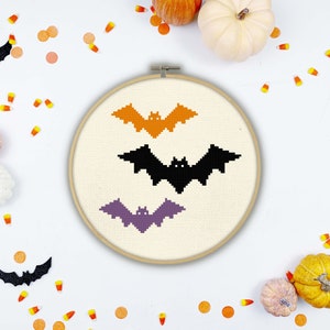 May include: A white embroidery hoop with three bats stitched on it. The bats are orange, black, and purple. The hoop is on a white background with scattered orange and yellow candy corn.