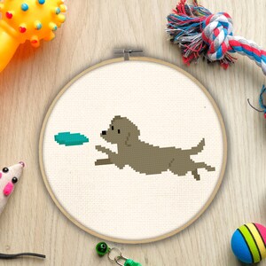 May include: A cross-stitch hoop art piece featuring a pixelated dog chasing a teal disc. The dog is brown and the background is off-white. The hoop is surrounded by dog toys, including a yellow bone, a rope toy, and a ball.