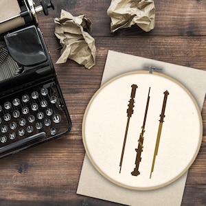 May include: A white embroidery hoop with three brown and gold wands inside. The hoop is on a wooden surface.