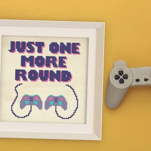 May include: Framed cross-stitch art with the text "JUST ONE MORE ROUND" in purple and blue. The artwork features two pixelated game controllers in the bottom center. A grey game controller is partially visible on the right side of the image.