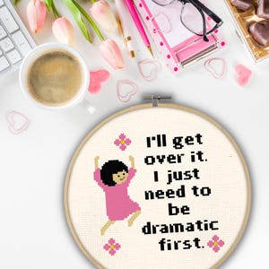 Op de afbeelding: Een witte borduurring met een roze en zwart kruissteekontwerp. Het ontwerp toont een cartoonfiguur dat springt met de tekst "I'll get over it. I just need to be dramatic first."