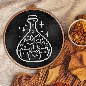 Könnte beinhalten: Schwarze und weiße Kreuzstich-Stickerei einer Zaubertrankflasche, die mit Katzen gefüllt ist. Die Flasche ist von Sternen umgeben.