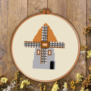 May include: A cross-stitch embroidery of a windmill in a wooden hoop. The windmill features a gray body, orange roof, and brown blades. The background is a cream-colored fabric with a wooden frame.