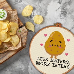 May include: A cross-stitch embroidery hoop with a cartoon potato with a cute face and the text "Less Haters, More Taters".
