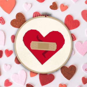 May include: A red heart with a white bandage over a crack in the heart. The heart is in a white embroidery hoop.