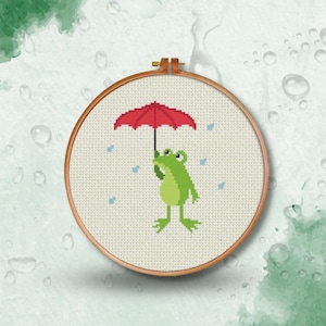 May include: A green frog holding a red umbrella with raindrops falling around it. The frog and umbrella are embroidered on a white fabric in a circular wooden hoop.