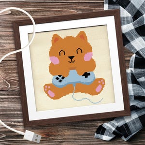 May include: Cross-stitch artwork in a brown frame. The image shows a brown bear character with pink cheeks and paws, holding a light blue video game controller. The bear is smiling. A white USB cable is in the foreground.