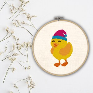 May include: A yellow chick wearing a pink and blue hat with a white background. The chick is embroidered on a white fabric stretched over a wooden embroidery hoop.