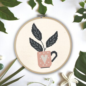 May include: A cross-stitch embroidery hoop featuring a plant design. The plant has dark gray leaves and is in a pink mug with a white heart. The background is a light beige fabric. The wooden hoop is surrounded by green leaves.