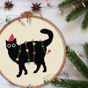 Puede incluir: Un bastidor de bordado de punto de cruz con un gato negro con un gorro de Papá Noel y luces navideñas. El gato está bordado sobre tela color crema, en un bastidor de madera. Decoración festiva navideña.