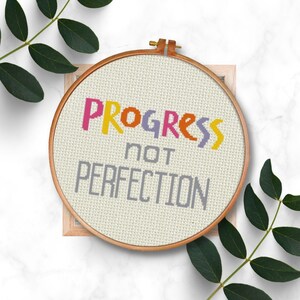 Könnte beinhalten: Ein weißer Stickrahmen mit einem beigefarbenen Stoffhintergrund. Der Stoff hat ein buntes Kreuzstich-Design mit der Aufschrift "PROGRESS not PERFECTION".
