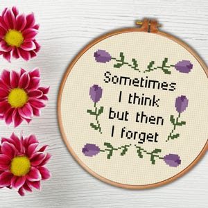 Puede incluir: Un bastidor de bordado de punto de cruz con el texto "Sometimes I think but then I forget" en negro. El diseño incluye flores moradas y detalles de hojas verdes. Tres flores rosas y amarillas están en el fondo.