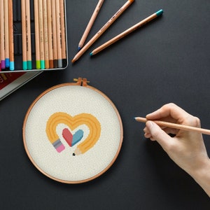 May include: A cross-stitch embroidery hoop with a design of two yellow pencils forming a heart shape with a red and blue heart inside.
