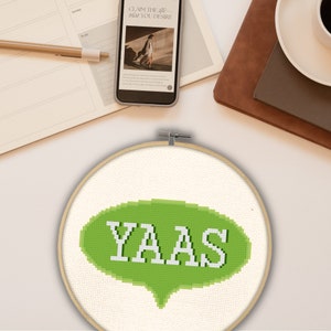 Puede incluir: Aro de bordado de punto de cruz verde y blanco con una burbuja de diálogo verde que dice "YAAS" en letras blancas.