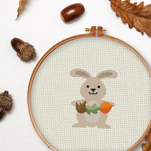 May include: A cross-stitch embroidery hoop with a beige background featuring a brown bunny holding a green leaf and an orange pumpkin. The bunny has a brown nose and black eyes.