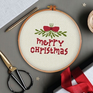 May include: A white embroidery hoop with a red and green cross-stitch design that says "merry christmas". The design includes a red bow and green leaves.