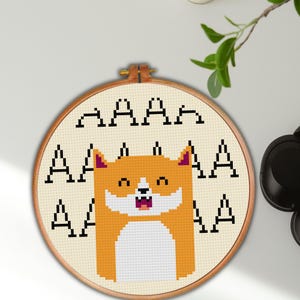 May include: A cross-stitch embroidery hoop featuring a cartoon Shiba Inu dog with an open mouth. The dog is orange and white, with black accents. The background is beige, and the text "AAAA" is stitched above the dog.