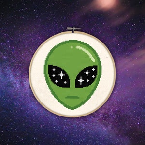 May include: A green alien face with black eyes and white stars in the eyes. The alien face is embroidered on a white fabric in a wooden embroidery hoop.
