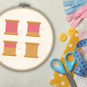May include: A white embroidery hoop with a cross-stitch design of four pink and brown thread spools. The hoop is surrounded by sewing supplies, including scissors, buttons, fabric scraps, and a measuring tape.