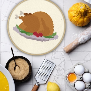 May include: A cross-stitch embroidery hoop featuring a pixelated roasted turkey on a platter. The image is surrounded by baking ingredients, including a small gourd, rolling pin, eggs, sugar, and a grater, suggesting a Thanksgiving theme.