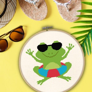 May include: A green frog wearing sunglasses and a red and blue inner tube, cross-stitched on white fabric in a wooden embroidery hoop.