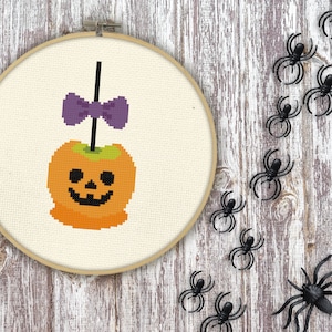 Puede incluir: Un patrón de punto de cruz de una manzana de caramelo de Halloween con un lazo morado en un palo. La manzana es naranja con una cara de calabaza. El fondo es una tela blanca.