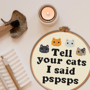 Puede incluir: Un bastidor de punto de cruz con fondo crema presenta cuatro caras de gato pixeladas y el texto "Tell your cats I said pspsps". El bastidor está enmarcado en madera. Una vela encendida y una hoja decorativa están en el fondo.