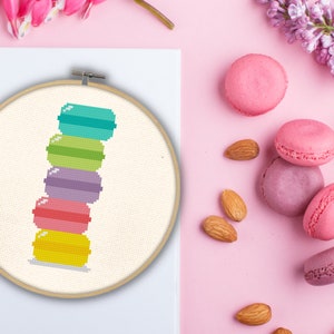 Op de afbeelding: Een stapel van zes kleurrijke macarons in een houten borduurring. De macarons zijn gerangschikt van boven naar beneden in de volgende volgorde: turquoise, groen, paars, roze, geel en wit.