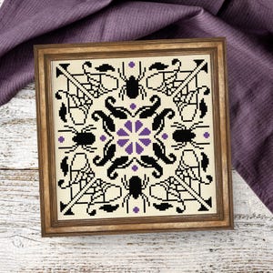 May include: A square framed cross-stitch embroidery featuring a symmetrical pattern on a cream-colored fabric. The design includes four black spiders, intricate black web motifs, and a central purple floral element with small purple accents.