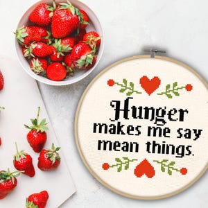 May include: A white embroidery hoop with a beige fabric background. The fabric has a black embroidered quote that reads "Hunger makes me say mean things."  The quote is surrounded by red hearts and green leaves.