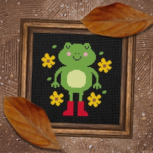 May include: Cross-stitch art of a green frog wearing red boots, surrounded by yellow flowers, set against a black background. The framed artwork has a gold-colored frame. Two brown leaves are placed on the top and bottom of the frame.