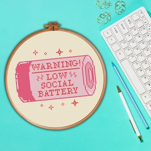 May include: A pink and white cross-stitch embroidery hoop with the text "WARNING! >> LOW >> SOCIAL BATTERY" in a pixelated font.