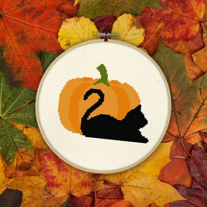 May include: A cross-stitch design featuring a black cat silhouette emerging from an orange pumpkin. The pumpkin has a green stem. The design is set in a wooden embroidery hoop, surrounded by autumn leaves.