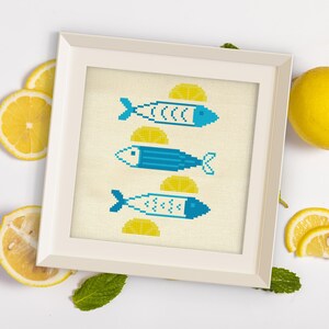 May include: A framed cross-stitch artwork featuring three stylized blue fish with yellow lemon slices. The artwork is in a white frame, surrounded by lemon slices and green leaves on a white background. The fish are in a pixelated style.