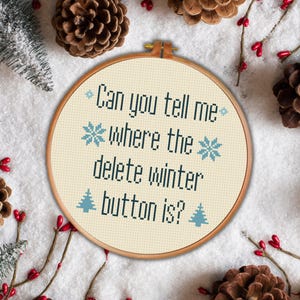 May include: A white embroidery hoop with a beige background and blue snowflakes and pine trees. The text reads "Can you tell me where the delete winter button is?"